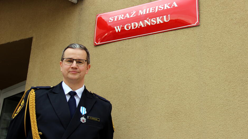 Na zdjęciu widać osobę w eleganckim, ciemnym mundurze służbowym stojącą przed budynkiem. Na mundurze widoczny jest odznaczeniowy medal przypięty do lewej strony klatki piersiowej. W tle znajduje się jasna ściana z czerwonym szyldem, na którym widnieje napis: „STRAŻ MIEJSKA W GDAŃSKU” Szyld jest prostokątny, z białymi literami na czerwonym tle, co wskazuje na oficjalne oznaczenie instytucji. Zdjęcie ma charakter formalny, prawdopodobnie wykonane w związku z uroczystością wręczenia odznaczeń lub wyróżnień.