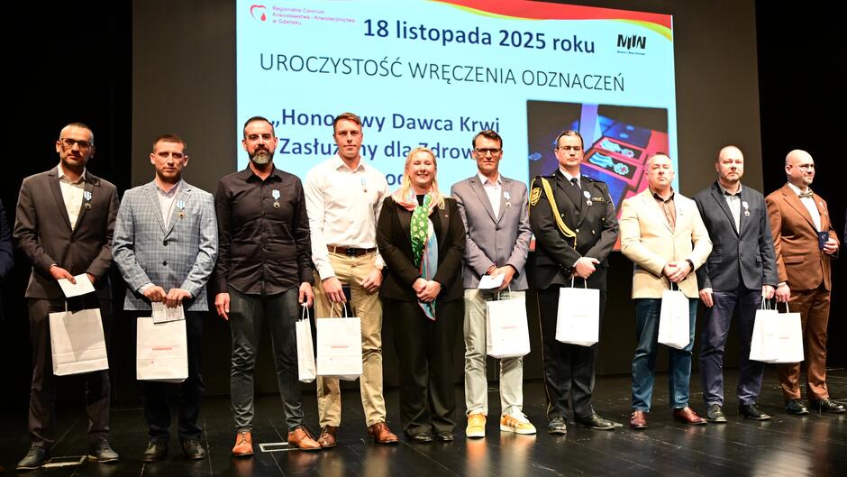 Na zdjęciu widać grupę osób stojących na scenie podczas oficjalnej uroczystości wręczenia odznaczeń. W tle znajduje się duży ekran z informacją o wydarzeniu: Data: 18 listopada 2025 roku Tytuł: „Uroczystość wręczenia odznaczeń” Treść: „Honorowy Dawca Krwi. Zasłużony dla zdrowia narodu”  Na ekranie widoczny jest również logotyp Narodowego Centrum Krwi oraz grafika przedstawiająca odznaczenia w pudełkach. Osoby na scenie trzymają w rękach białe torby prezentowe, a część z nich ma przypięte medale na marynarkach. Ubrania są eleganckie – garnitury, koszule, jedna osoba w mundurze służbowym. Scena jest dobrze oświetlona, a tło czarne, co podkreśla formalny charakter wydarzenia.