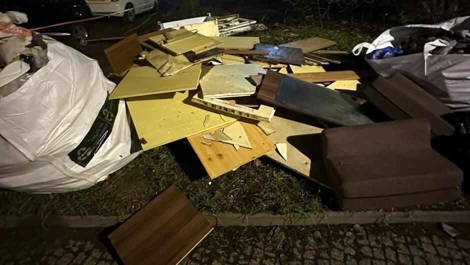 Na zdjęciu widać stertę odpadów wielkogabarytowych i budowlanych zgromadzonych na chodniku lub przy krawędzi jezdni. W skład tej sterty wchodzą: Płyty drewniane i laminowane – różnej wielkości, w tym elementy mebli (np. fronty szafek, półki).
Fragmenty tapicerowanych mebli – widoczny jest co najmniej jeden element przypominający część kanapy lub fotela w kolorze brązowym.
Worki typu Big Bag – duże białe worki na odpady, częściowo wypełnione.
Metalowe elementy – możliwe fragmenty konstrukcji lub okuć.
Tło – widać zaparkowane samochody i fragment ulicy, zdjęcie wykonane nocą przy sztucznym oświetleniu.

Całość sprawia wrażenie nielegalnego składowiska lub miejsca tymczasowego gromadzenia odpadów po remoncie lub likwidacji wyposażenia mieszkania.
