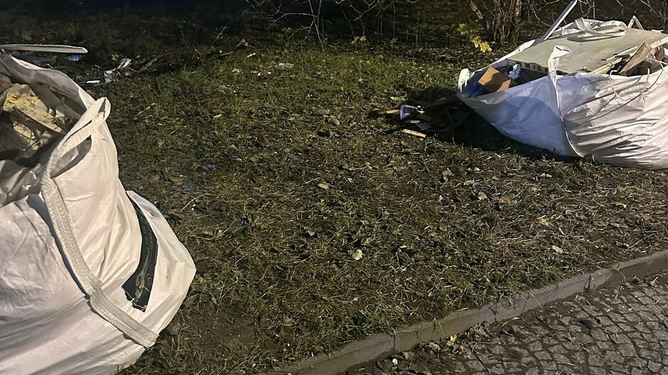 Na zdjęciu widać fragment trawnika przy krawężniku, obok chodnika z kostki brukowej. Na trawie znajdują się dwa duże białe worki typu Big Bag, wypełnione odpadami budowlanymi lub remontowymi – widać m.in. kawałki drewna, płyt, gruzu i innych materiałów. Worki są częściowo otwarte, a wokół nich leżą drobne śmieci. W tle widoczna jest zieleń – krzewy i gałęzie – w słabym oświetleniu, co sugeruje, że zdjęcie zostało wykonane wieczorem lub w nocy.