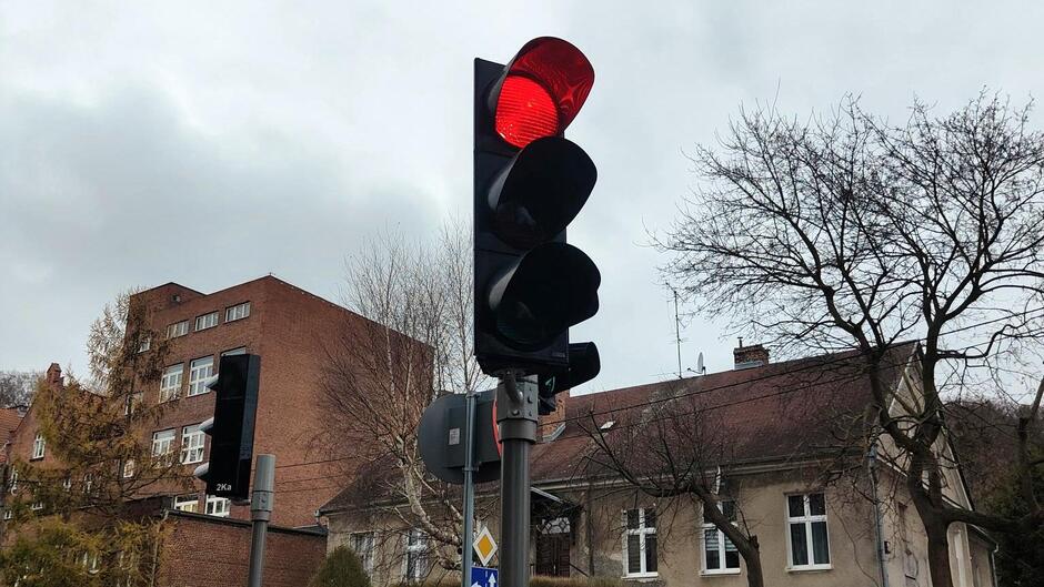 Na zdjęciu widać nowo zamontowany sygnalizator świetlny na słupie przy skrzyżowaniu. Urządzenie jest w pełni sprawne – świeci się czerwone światło. W tle znajdują się budynki mieszkalne z cegły i tynkowane, bez liści na drzewach, co wskazuje na późną jesień. Niebo jest pochmurne, w odcieniach szarości, co nadaje zdjęciu stonowany klimat.
To zdjęcie dobrze pokazuje efekt naprawy – sygnalizator działa i jest na swoim miejscu.