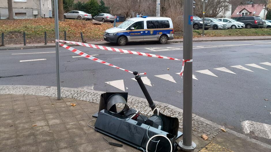 Na zdjęciu widać uszkodzony sygnalizator świetlny leżący na chodniku, tuż obok słupa, na którym był zamontowany. Urządzenie jest rozbite – widać główną część z soczewkami i przewodami oraz oderwane ramię mocujące. Całość została zabezpieczona biało-czerwoną taśmą ostrzegawczą, rozpiętą pomiędzy słupami.
W tle znajduje się jezdnia z oznaczeniami przejścia dla pieszych oraz zaparkowany pojazd Straży Miejskiej z włączonymi światłami ostrzegawczymi. Dalej widoczne są samochody na parkingu, fragmenty budynków i opadłe liście na trawniku, co sugeruje późną jesień.