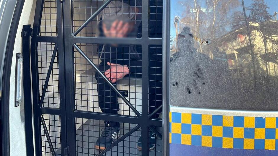 Na zdjęciu widać wnętrze przedziału transportowego w radiowozie Straży Miejskiej, zabezpieczone metalową kratą. W środku znajduje się osoba siedząca na ławce, z rękami opuszczonymi w dół. Widoczne są sportowe buty oraz ciemne ubranie. Po prawej stronie kadru widać fragment zewnętrznej części pojazdu z charakterystycznym niebiesko-żółtym wzorem oznaczeń służb miejskich. W tle, za szybą, odbijają się drzewa i zabudowania, co sugeruje, że scena ma miejsce na zewnątrz, w ciągu dnia.