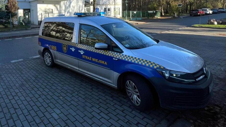 Na zdjęciu widoczny jest radiowóz Straży Miejskiej zaparkowany na utwardzonej nawierzchni z kostki brukowej. Pojazd ma charakterystyczne oznaczenia służb – niebiesko-żółtą szachownicę na bokach, napis „STRAŻ MIEJSKA” oraz numer alarmowy 986. Na dachu znajduje się niebieski sygnał świetlny. W tle widać fragment ulicy, ogrodzenie z siatki, drzewa oraz budynki, co sugeruje, że scena ma miejsce w spokojnej, miejskiej okolicy w ciągu dnia.