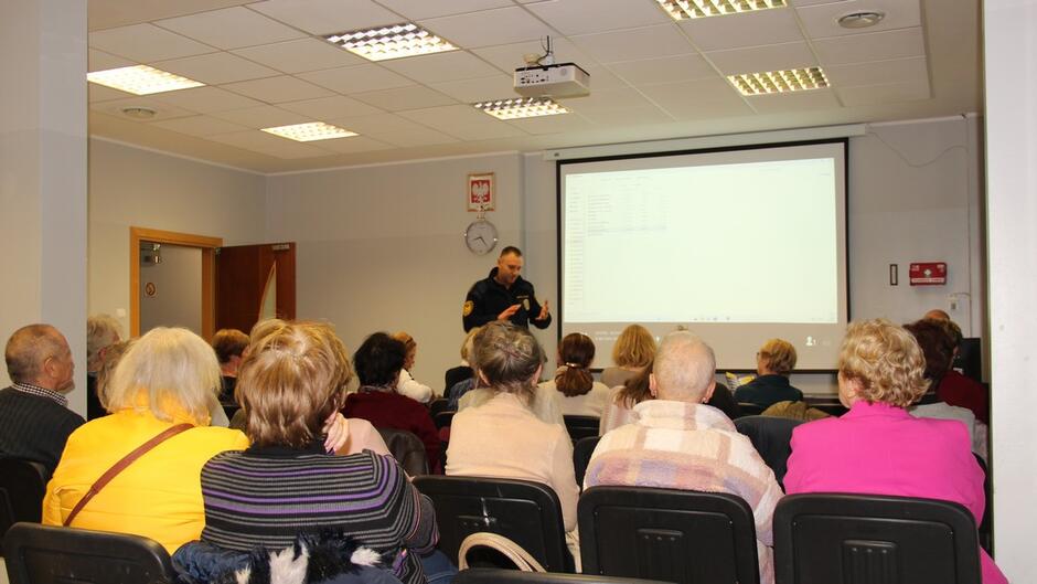 Sala konferencyjna z rzędem krzeseł ustawionych w kierunku ekranu projekcyjnego. Na ekranie wyświetlony jest interfejs komputera z listą plików. W pierwszym planie widać grupę osób siedzących tyłem, uważnie słuchających prelegenta stojącego przy ekranie. Na suficie zamontowany jest projektor, a na ścianie zegar i tabliczka z godłem Polski. Oświetlenie jest jasne, typowe dla sali szkoleniowej.