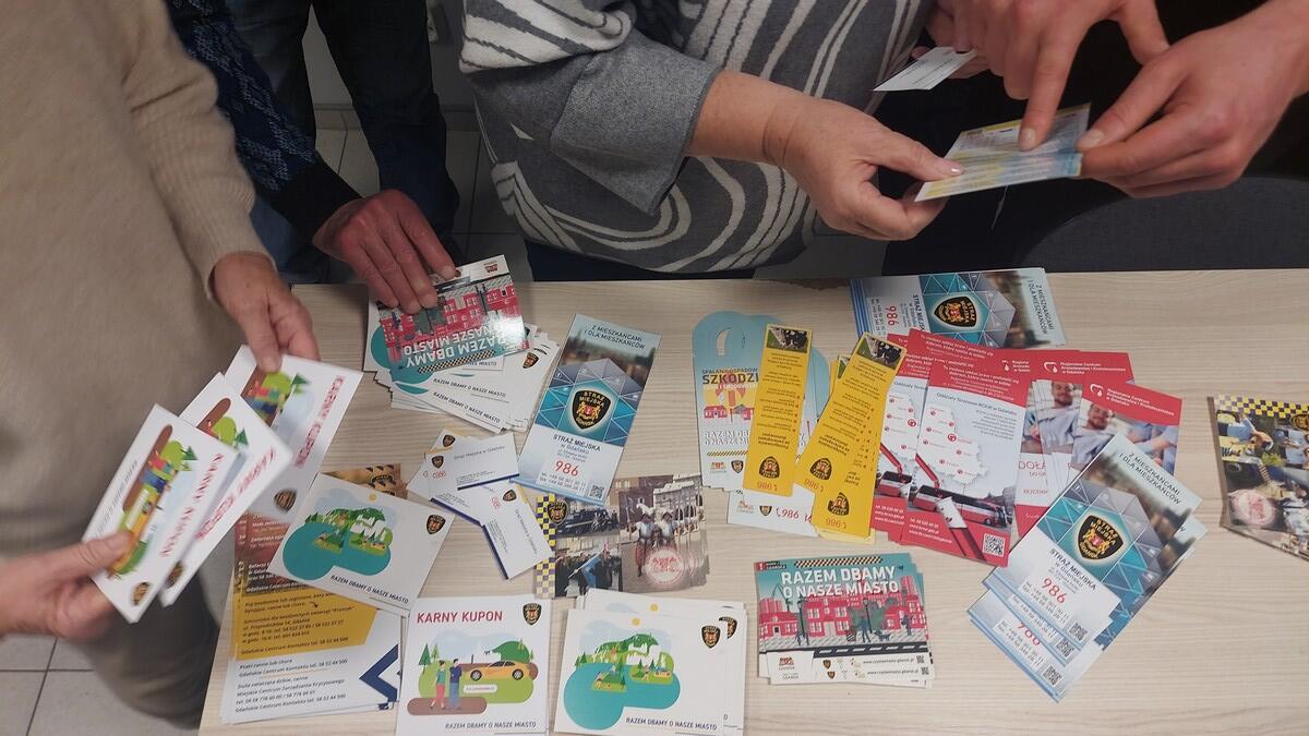 Kilka osób stoi przy stole, na którym rozłożone są kolorowe ulotki i broszury informacyjne. Materiały dotyczą różnych tematów związanych z bezpieczeństwem, w tym ostrzeżeń przed oszustwami i kontaktów do służb. Na ulotkach widoczne są grafiki, logotypy oraz hasła edukacyjne. Uczestnicy przeglądają materiały, trzymając je w dłoniach.