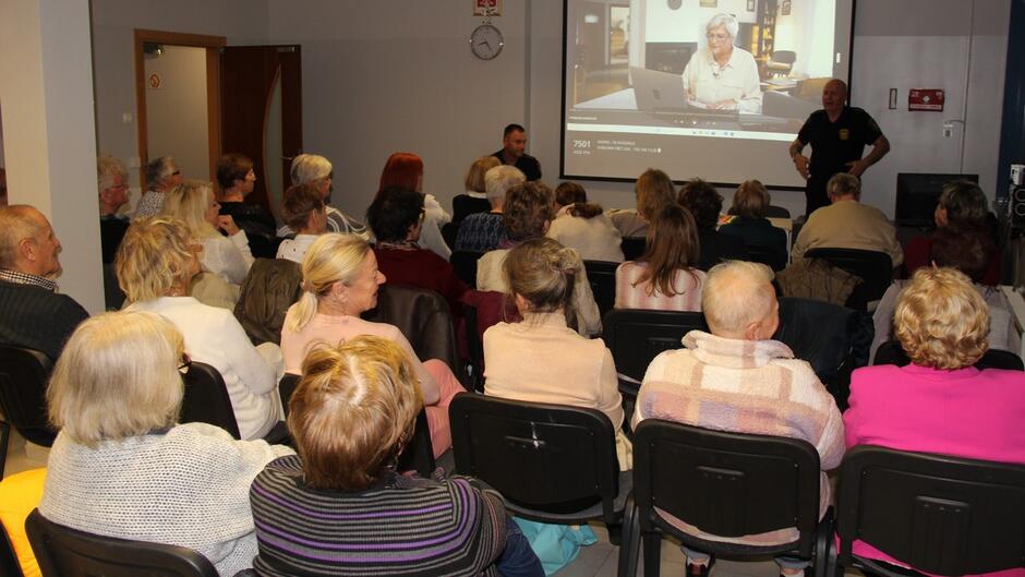 Sala szkoleniowa z uczestnikami siedzącymi w rzędach, zwróconymi w stronę ekranu. Na ekranie wyświetlony jest film przedstawiający starszego mężczyznę przy komputerze, co sugeruje temat związany z cyberbezpieczeństwem lub oszustwami internetowymi. Prelegent stoi obok ekranu, a w tle widać drzwi i zegar na ścianie.