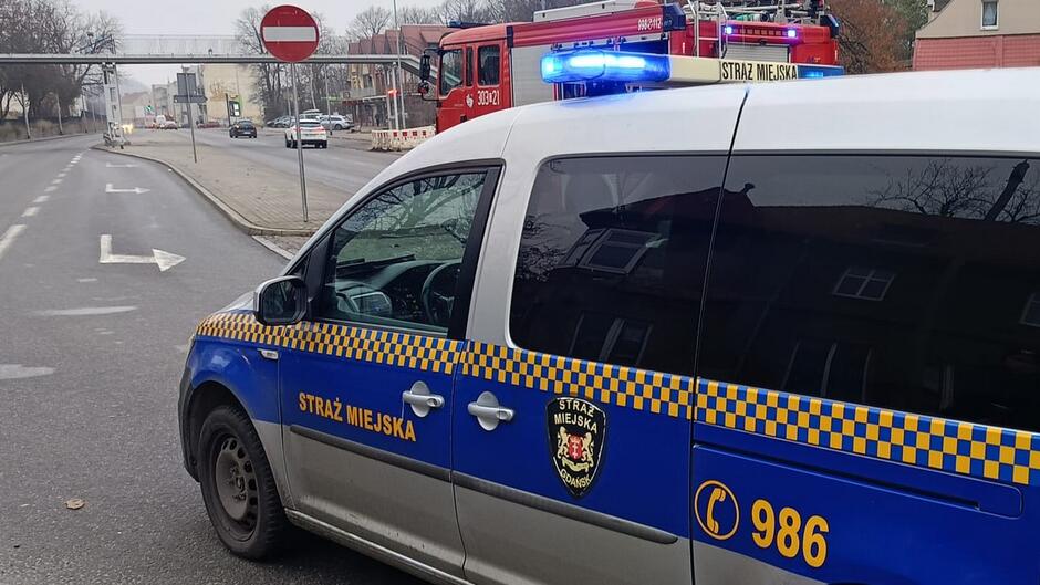 Samochód Straży Miejskiej w Gdańsku (biało-niebieski pojazd z żółtymi napisami „STRAŻ MIEJSKA”) stoi na ulicy z włączonymi niebieskimi światłami ostrzegawczymi.
Dodatkowe szczegóły: W tle widoczny jest wóz strażacki oraz fragment ulicy z oznakowaniem „zakaz wjazdu”.
Tło: Pusta jezdnia, zabudowa miejska, zimowa aura (brak liści na drzewach).