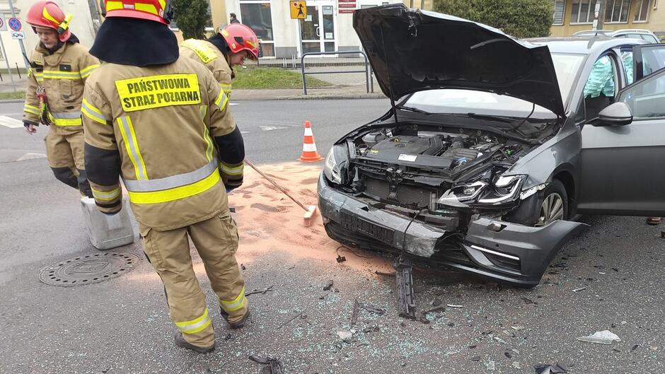Widok szerszy na miejsce wypadku. Widać ten sam pojazd (ciemnoszary hatchback) na środku skrzyżowania, otoczony przez strażaków i ratowników.
Dodatkowe szczegóły: Na drodze ustawione są pomarańczowe pachołki ostrzegawcze. Z tyłu stoi czerwony wóz strażacki z oznaczeniem „303[21]”.
Tło: Budynki mieszkalne, chodniki, kilka osób obserwujących sytuację. Droga jest częściowo zablokowana.