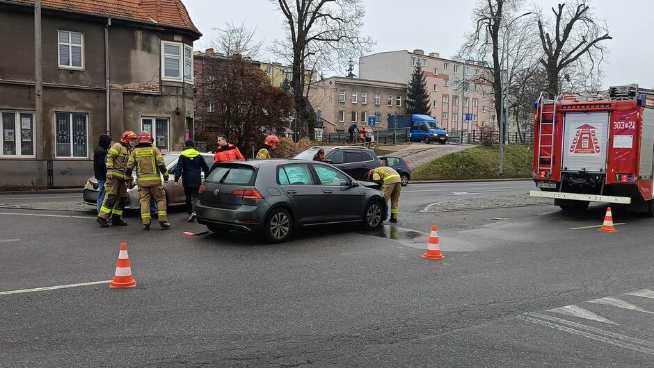 Widać samochód osobowy w kolorze ciemnoszarym, który brał udział w wypadku drogowym. Auto ma poważnie uszkodzony przód – zderzak jest oderwany, maska podniesiona, a elementy silnika są odsłonięte.
Dodatkowe szczegóły: Na jezdni leży rozbite szkło i fragmenty plastikowych części pojazdu. Widać rozsypany sorbent (substancja do neutralizacji płynów) w okolicy przodu auta.
Osoby: Strażacy w pełnym umundurowaniu z napisem „PAŃSTWOWA STRAŻ POŻARNA” na plecach pracują przy zabezpieczaniu miejsca zdarzenia. Jeden z nich trzyma pojemnik, prawdopodobnie z sorbentem.
Tło: Ulica miejska, chodnik, budynki mieszkalne i usługowe.