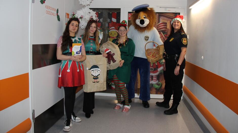 grupa przebrana za elfy i straznik w stroju lwa oraz strażniczka w czapce mikołaja stoją na korytarzy na tle ozdobionych świątecznie drzwi na oddział.