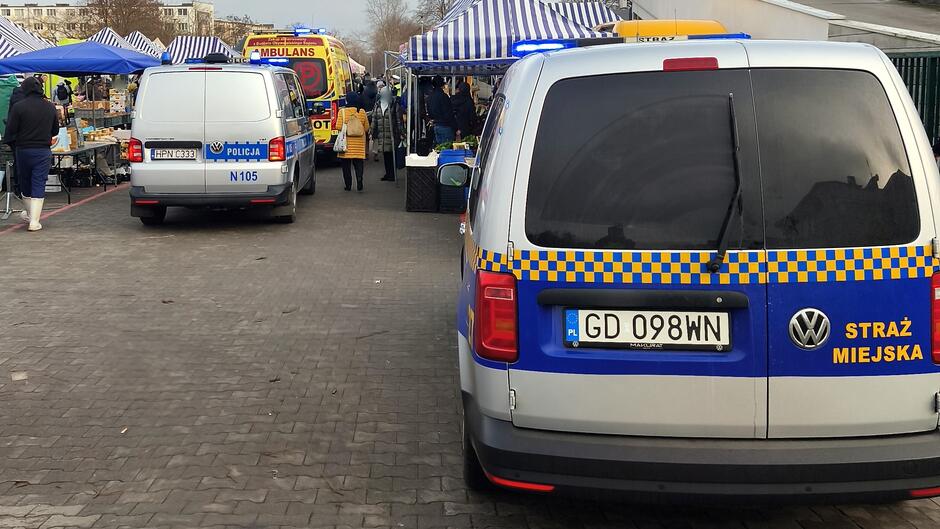 Na zdjęciu widać fragment targowiska z kilkoma pojazdami służb ratunkowych i porządkowych.
Opis szczegółowy: Pierwszy plan: Samochód Straży Miejskiej (Volkswagen) z widocznym napisem „STRAŻ MIEJSKA” na tylnej części pojazdu oraz tablicą rejestracyjną „GD 098WN”. Auto ma charakterystyczne niebiesko-żółte oznakowanie i koguta na dachu.
Środkowa część zdjęcia: Drugi pojazd – policyjny, oznakowany napisem „POLICJA” i tablicą rejestracyjną „HPN 88E1”. Obok niego widać osoby poruszające się po targowisku.
Tło: Ambulans z napisem „AMBULANS” i „RATOWNICTWO MEDYCZNE” na górze, z włączonymi światłami ostrzegawczymi. Wokół znajdują się stragany z niebiesko-białymi pasami na daszkach, a na stołach leżą różne towary.
Otoczenie: Nawierzchnia jest brukowana, a w tle widać zabudowania i drzewa bez liści, co sugeruje porę jesienno-zimową.
Cała scena wygląda na interwencję służb na terenie targowiska – obecność Straży Miejskiej, Policji i karetki wskazuje na zdarzenie wymagające ich obecności.