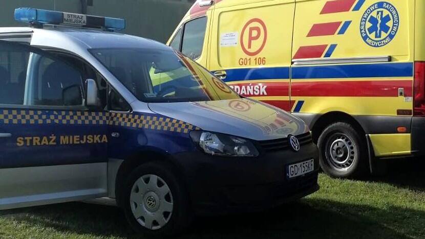 Na zdjęciu widać dwa pojazdy służb ratunkowych stojące na trawie:
Pierwszy plan: Radiowóz Straży Miejskiej marki Volkswagen. Samochód jest oznakowany charakterystycznymi barwami – granatowy dół, srebrna góra, żółto-niebieska szachownica na bokach oraz napis „STRAŻ MIEJSKA”. Na dachu znajduje się niebieski kogut sygnalizacyjny. Widoczna jest tablica rejestracyjna zaczynająca się od „GD”.
Tło: Karetka pogotowia ratunkowego w barwach żółto-czerwonych z niebieskimi elementami. Na boku pojazdu widoczny jest napis „Państwowe Ratownictwo Medyczne”, symbol gwiazdy życia oraz oznaczenie „Gdańsk”. Na drzwiach znajduje się czerwone kółko z literą „P” i numerem „G01 106”.
Otoczenie: Pojazdy stoją na zielonej trawie, w tle widać fragment budynku lub ogrodzenia w neutralnym kolorze.
Cała scena wygląda na sytuację, w której Straż Miejska i pogotowie ratunkowe współpracują w ramach interwencji lub zabezpieczenia wydarzenia.