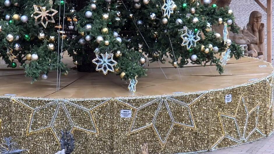 Na zdjęciu znajduje się dolna część dużej, świątecznej choinki ustawionej na podwyższonej konstrukcji. Choinka jest bogato udekorowana bombkami w kolorach złotym, srebrnym i zielonym oraz ozdobami w kształcie gwiazd. Podstawę drzewka otacza dekoracyjna osłona w złotym kolorze, wykonana z błyszczących elementów, dodatkowo ozdobiona dużymi wzorami gwiazd z jasnych lampek.
Na osłonie widoczne są tabliczki ostrzegawcze z napisem informującym o zakazie wchodzenia i siadania na konstrukcji. Całość znajduje się na tle brukowanego placu, a w tle widać fragment drewnianej zabudowy, prawdopodobnie elementy jarmarku świątecznego.
Świąteczne dekoracje i złote akcenty nadają zdjęciu elegancki, bożonarodzeniowy charakter.