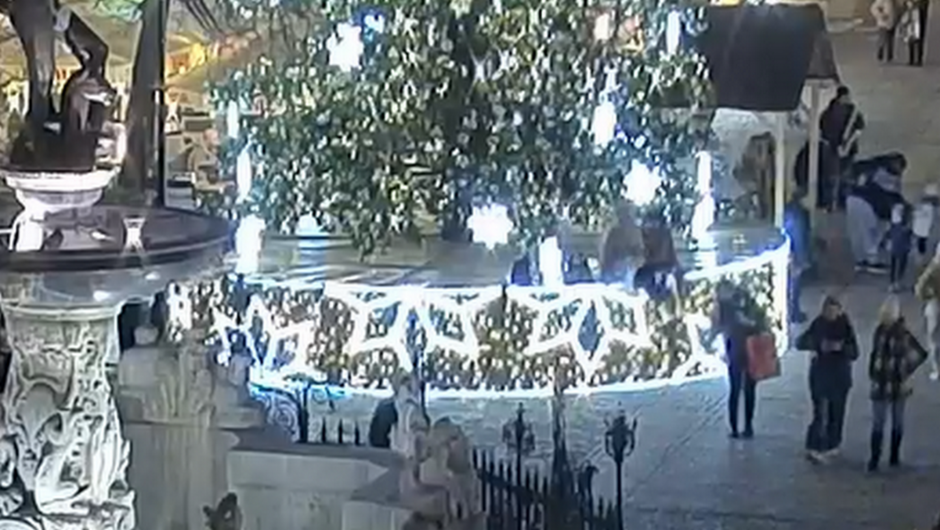 Na zdjęciu widać fragment Długiego Targu w Gdańsku, udekorowany na okres świąteczny. Centralnym elementem jest duża, bogato oświetlona choinka z licznymi ozdobami i jasnymi dekoracjami w kształcie gwiazdek. Choinka stoi na podwyższonej konstrukcji, której osłona jest ozdobiona światełkami i złotymi elementami, tworząc efektowną iluminację.
Na pierwszym planie, po lewej stronie, widoczna jest część Fontanny Neptuna – charakterystycznego zabytku Gdańska. Wokół choinki znajduje się kilka osób, część z nich stoi obok, a dwie osoby są widoczne na osłonie balastu, mimo że obowiązuje zakaz wchodzenia na tę konstrukcję.
Tło zdjęcia pokazuje fragment zabudowy Długiego Targu oraz przechodniów spacerujących w świątecznej scenerii. Całość jest dobrze oświetlona, co podkreśla atmosferę jarmarku bożonarodzeniowego.