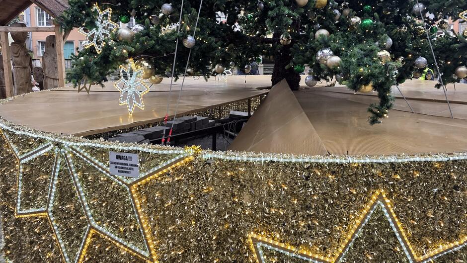 Na zdjęciu widać dolną część dużej, świątecznej choinki ustawionej na podwyższonej konstrukcji. Choinka jest bogato udekorowana bombkami w kolorach złotym, srebrnym i zielonym oraz ozdobami w kształcie gwiazd. Podstawę drzewka otacza dekoracyjna osłona w złotym kolorze, wykonana z błyszczących elementów, dodatkowo ozdobiona dużymi wzorami gwiazd z jasnych lampek.
Na osłonie znajduje się wyraźna tabliczka ostrzegawcza z napisem:
UWAGA !!!
ZAKAZ WCHODZENIA, SIADANIA
NA OSŁONIE BALASTU,
MASKOWNICY Widać również, że część osłony jest uszkodzona – jej powierzchnia zapadła się, odsłaniając elementy konstrukcji pod spodem. To pokazuje, jak niebezpieczne może być wchodzenie na tę część instalacji.
Tło zdjęcia przedstawia fragment zabudowy Długiego Targu oraz drewniane elementy dekoracyjne, prawdopodobnie związane z jarmarkiem świątecznym.