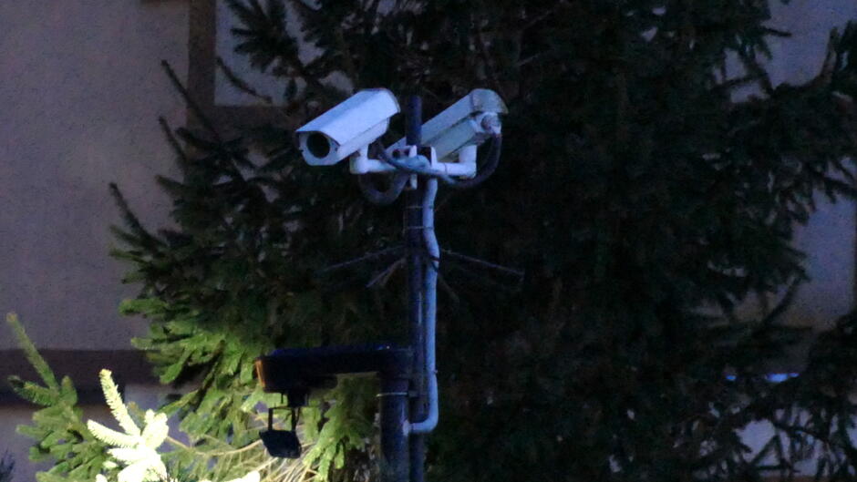 Na zdjęciu widać zestaw kamer monitoringu zamontowanych na metalowym słupie. Na górze znajdują się dwie białe kamery skierowane w różne strony, co sugeruje, że mają za zadanie obserwować szeroki obszar. Poniżej znajduje się dodatkowe urządzenie – prawdopodobnie reflektor lub czujnik, który może wspomagać działanie kamer w warunkach słabego oświetlenia. W tle widoczne są gęste gałęzie drzew iglastych oraz fragment elewacji budynku w kolorze jasnobrązowym. Zdjęcie zostało wykonane w warunkach słabego światła, prawdopodobnie wieczorem lub nocą, co podkreśla znaczenie monitoringu w zapewnianiu bezpieczeństwa.