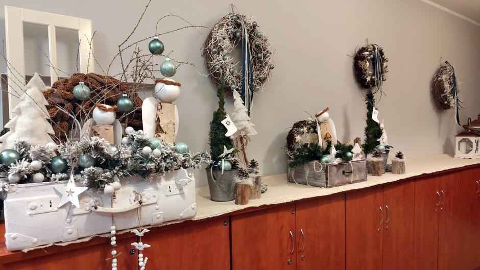 Na zdjęciu widać elegancką, świąteczną aranżację ustawioną na długim blacie z drewnianymi szafkami. Dekoracje utrzymane są w naturalnym, zimowym stylu – dominują odcienie bieli, srebra i zieleni. Na blacie znajdują się różnorodne kompozycje: ozdobne wianki z gałązek, małe choinki, gałązki iglaste, szyszki, bombki w kolorze srebrnym i turkusowym, a także elementy z drewna i bawełny imitujące śnieg. Niektóre dekoracje umieszczone są w metalowych wiaderkach, drewnianych skrzynkach lub na białych podstawach, co nadaje całości rustykalny charakter. W tle na ścianie wiszą kolejne wianki, a całość jest starannie ułożona, tworząc przytulny, świąteczny klimat.