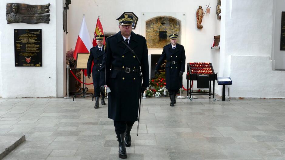 Na zdjęciu widać trzech funkcjonariuszy w ciemnych, galowych mundurach, pełniących wartę honorową wewnątrz świątyni. Dwóch z nich stoi w tle, a jeden znajduje się na pierwszym planie, idąc w kierunku aparatu. Funkcjonariusze mają na głowach czapki służbowe, a jeden z nich trzyma szablę. W tle widoczna jest ściana z jasnego tynku, na której znajdują się tablice pamiątkowe oraz polska flaga i sztandar. Centralnie umieszczony jest grób lub miejsce pamięci, udekorowane kwiatami i wieńcami. Po prawej stronie znajduje się stolik z zapalonymi świecami w czerwonych osłonkach, a obok skrzynka na ofiary. Całość odbywa się w przestrzeni o kamiennej posadzce, co sugeruje wnętrze zabytkowej bazyliki.
