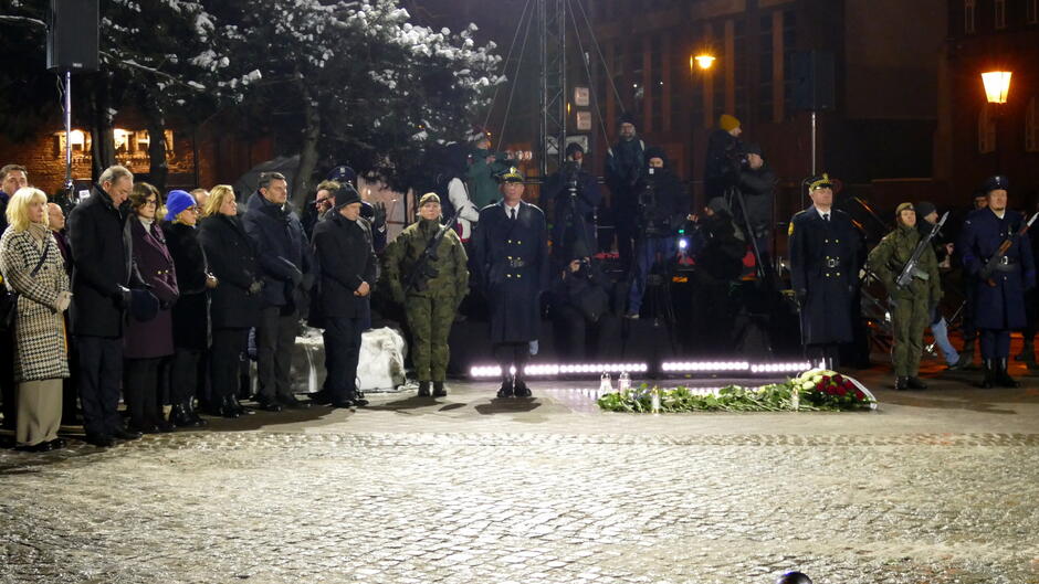 Na zdjęciu widać uroczystość odbywającą się na otwartym powietrzu, prawdopodobnie w godzinach wieczornych lub nocnych, co sugeruje ciemne tło i sztuczne oświetlenie.
Opis szczegółowy: W centralnej części znajduje się grupa żołnierzy w galowych mundurach oraz w umundurowaniu polowym, stojących w pozycji honorowej.
Przed nimi, na bruku, ułożone są wieńce i wiązanki kwiatów, co wskazuje na charakter upamiętniający lub ceremonialny.
Po lewej stronie stoi grupa osób w eleganckich płaszczach i zimowych ubraniach, co sugeruje chłodną porę roku.
W tle widoczne są elementy dekoracyjne, oświetlenie oraz drzewo pokryte śniegiem, co dodatkowo potwierdza zimową scenerię.
Oświetlenie jest intensywne, skierowane na uczestników uroczystości, a w tle widać latarnie uliczne i fragmenty zabudowy miejskiej.

Całość przedstawia podniosłą atmosferę ceremonii patriotycznej lub rocznicowej, z udziałem wojska i przedstawicieli społeczności.