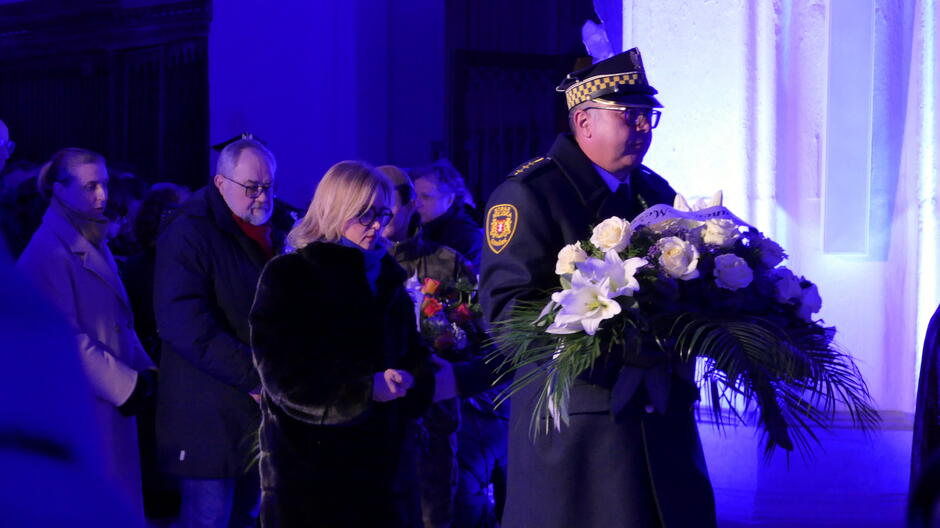 Na zdjęciu widać scenę z uroczystości odbywającej się w pomieszczeniu o jasnych ścianach, oświetlonym intensywnym, niebieskim światłem.
Opis szczegółowy: Na pierwszym planie znajduje się funkcjonariusz w ciemnym, galowym mundurze z czapką służbową, trzymający okazały wieniec z białymi kwiatami (m.in. lilie i róże) oraz zielonymi liśćmi.
Za nim widoczna jest grupa osób ubranych w zimowe płaszcze, trzymających kwiaty i wieńce, co sugeruje udział w ceremonii upamiętniającej.
Tło stanowi jasna ściana z pionowym elementem architektonicznym, podświetlona na niebiesko, co nadaje scenie podniosły charakter.
Całość wskazuje na uroczystość o charakterze patriotycznym lub żałobnym, odbywającą się w eleganckim, zamkniętym wnętrzu.