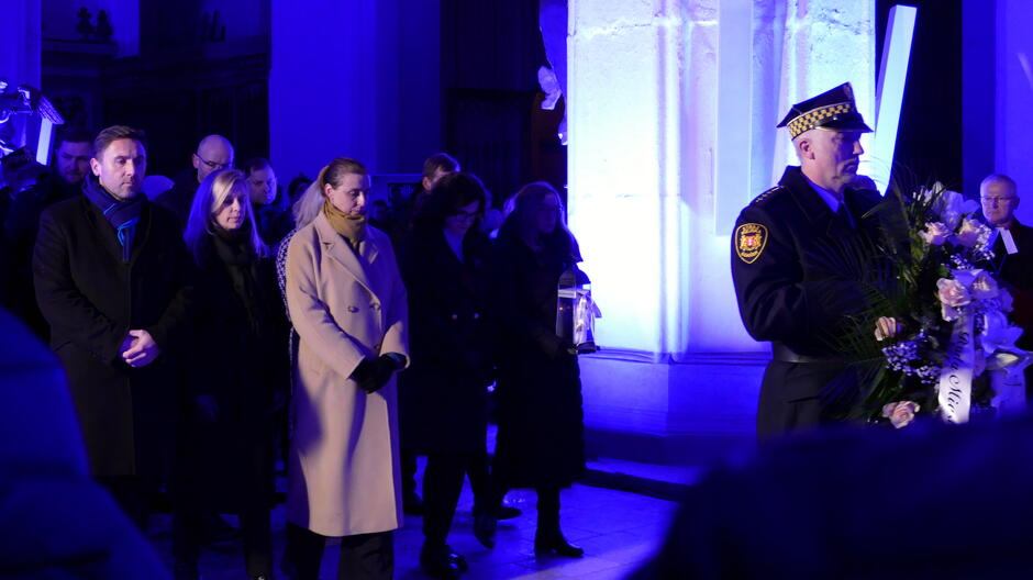 Na zdjęciu widać uroczystość odbywającą się w eleganckim wnętrzu, oświetlonym intensywnym, niebieskim światłem, co nadaje scenie podniosły charakter. Opis szczegółowy: Na pierwszym planie znajduje się funkcjonariusz w galowym mundurze z czapką służbową, trzymający okazały wieniec z białymi i jasnoróżowymi kwiatami oraz zielonymi liśćmi. Na wieńcu widoczna jest biało-czarna szarfa z napisem. W tle stoi grupa osób w eleganckich płaszczach, ustawionych w rzędzie, co sugeruje udział w ceremonii upamiętniającej lub patriotycznej. Tło stanowi jasna ściana z pionowym elementem architektonicznym, podświetlona na niebiesko, a cała scena odbywa się w przestrzeni o charakterze sakralnym lub zabytkowym. Oświetlenie jest mocne, skierowane na uczestników, co podkreśla uroczysty nastrój wydarzenia.