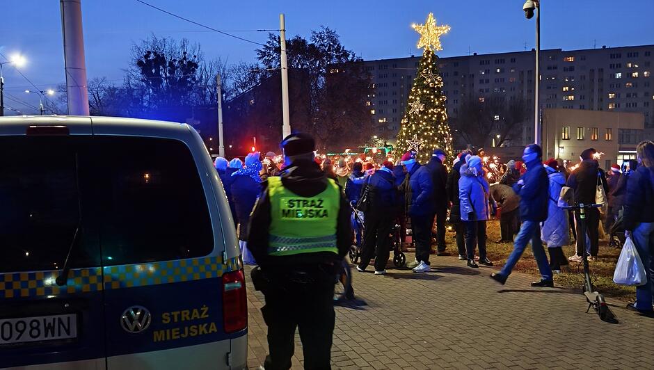 Na zdjęciu widać strażnika miejskiego stojącego obok oznakowanego radiowozu Straży Miejskiej. W tle zgromadziła się grupa osób uczestniczących w świątecznym wydarzeniu. Centralnym punktem jest duża, oświetlona choinka z gwiazdą na szczycie. Scena rozgrywa się po zmroku, a okolicę rozświetlają kolorowe lampki i dekoracje.