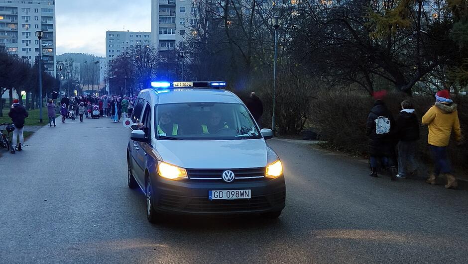 Na zdjęciu widać oznakowany radiowóz Straży Miejskiej z włączonymi niebieskimi światłami, jadący alejką osiedlową. W tle znajduje się grupa osób uczestniczących w wydarzeniu lub przemarszu, a po bokach alei idą kolejne osoby w kierunku zgromadzenia. Scena odbywa się o zmierzchu, w otoczeniu drzew i wysokich bloków mieszkalnych.