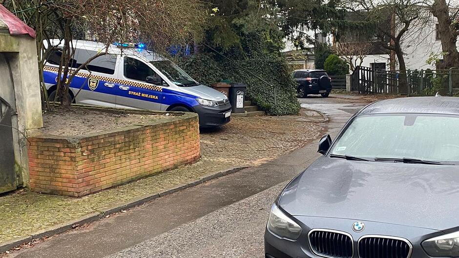 Na zdjęciu widać uliczkę osiedlową otoczoną drzewami i zabudową jednorodzinną. Po lewej stronie stoi oznakowany radiowóz Straży Miejskiej z włączonymi światłami, zaparkowany przy murku z cegły. Na pierwszym planie, po prawej stronie, znajduje się zaparkowany samochód osobowy. W głębi kadru widoczny jest zakręt prowadzący do kolejnych posesji oraz kilka innych pojazdów.