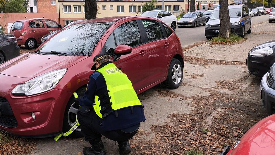 Na zdjęciu widać funkcjonariusza Straży Miejskiej w żółtej kamizelce odblaskowej, który zakłada blokadę na koło czerwonego samochodu osobowego zaparkowanego na poboczu. W tle znajduje się ulica z zaparkowanymi pojazdami oraz budynki mieszkalne. Na chodniku i jezdni leżą jesienne liście.
