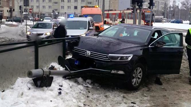 Na zdjęciu widać czarny samochód osobowy, który uderzył w barierki oddzielające przystanek od jezdni. Z przodu pojazdu leży przewrócony sygnalizator świetlny. Na miejscu są służby — widoczne są radiowóz, pojazdy techniczne oraz kilku funkcjonariuszy. Na ulicy panują zimowe warunki, a w tle stoją tramwaje i inne samochody.