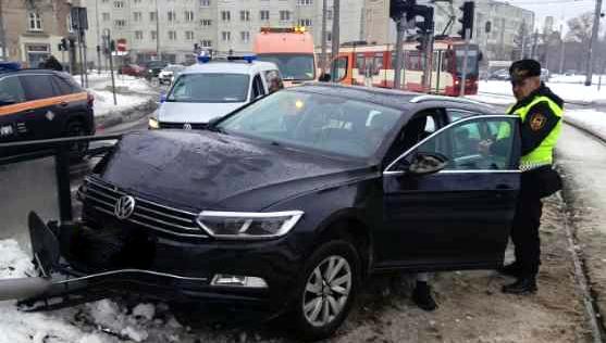 Na zdjęciu widać czarny samochód osobowy, który uderzył w barierki przy przystanku tramwajowym. Uszkodzony sygnalizator świetlny leży przed przodem pojazdu. Obok auta stoi funkcjonariusz w żółtej kamizelce, a w tle widoczne są radiowozy, pojazd techniczny oraz tramwaj. Na ulicy panują zimowe warunki — jezdnia i chodniki są pokryte śniegiem.