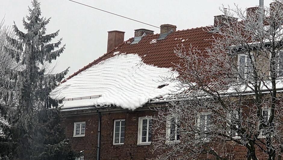 Na dachu budynku mieszkalnego zalega duża warstwa zmarzniętego śniegu, która częściowo osunęła się w stronę krawędzi. Śnieg tworzy wyraźne fale i zwisy nad linią rynny, stwarzając potencjalne zagrożenie dla przechodniów. Wokół widać oszronione drzewa, a całość utrzymana jest w zimowej, mroźnej scenerii.