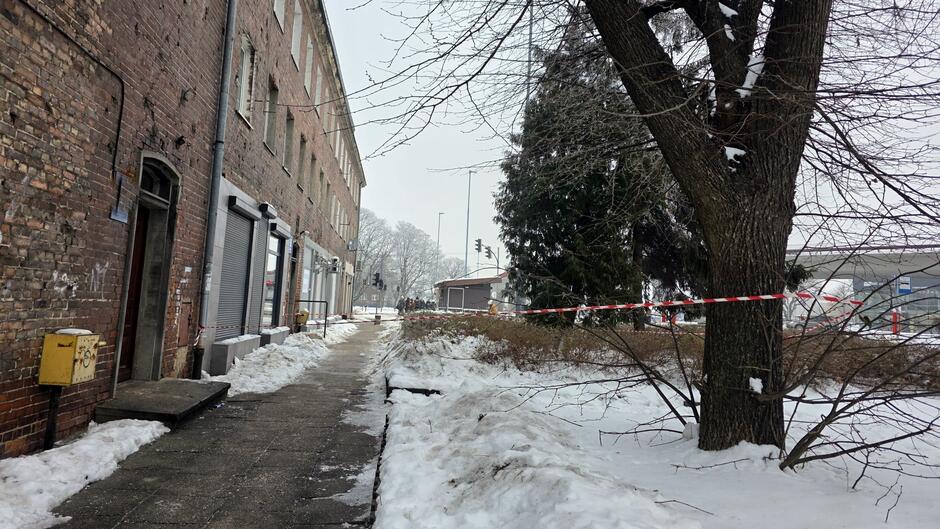 Zimowa sceneria przy budynku mieszkalnym. Fragment chodnika został odgrodzony biało‑czerwoną taśmą ze względu na zagrożenie spadającym śniegiem z dachu. Na ziemi widać zalegający śnieg, a pobliskie drzewa i krzewy pokryte są szronem. W głębi kadru widoczne są kolejne budynki oraz sygnalizacja świetlna.