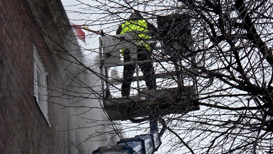 Pracownik służb technicznych usuwa zalegający śnieg z krawędzi dachu, stojąc na podnośniku koszowym. Osoba w jaskrawej kamizelce pracuje na wysokości, a wokół widać gałęzie drzew i opadający śnieg usuwany z powierzchni budynku.