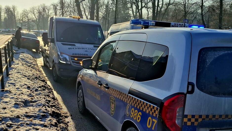 Na pierwszym zdjęciu widać sytuację drogową z udziałem służb. Na pierwszym planie stoi samochód oznakowany jako pojazd Straży Miejskiej z włączonymi światłami na dachu. Przed nim znajduje się laweta, która podjechała prawdopodobnie po uszkodzony samochód widoczny dalej. Na poboczu, w śniegu, stoi rozbity pojazd, przy którym znajduje się jedna osoba. Droga jest częściowo oblodzona i ośnieżona, a w tle widać drzewa bez liści – wygląda na zimowy poranek.