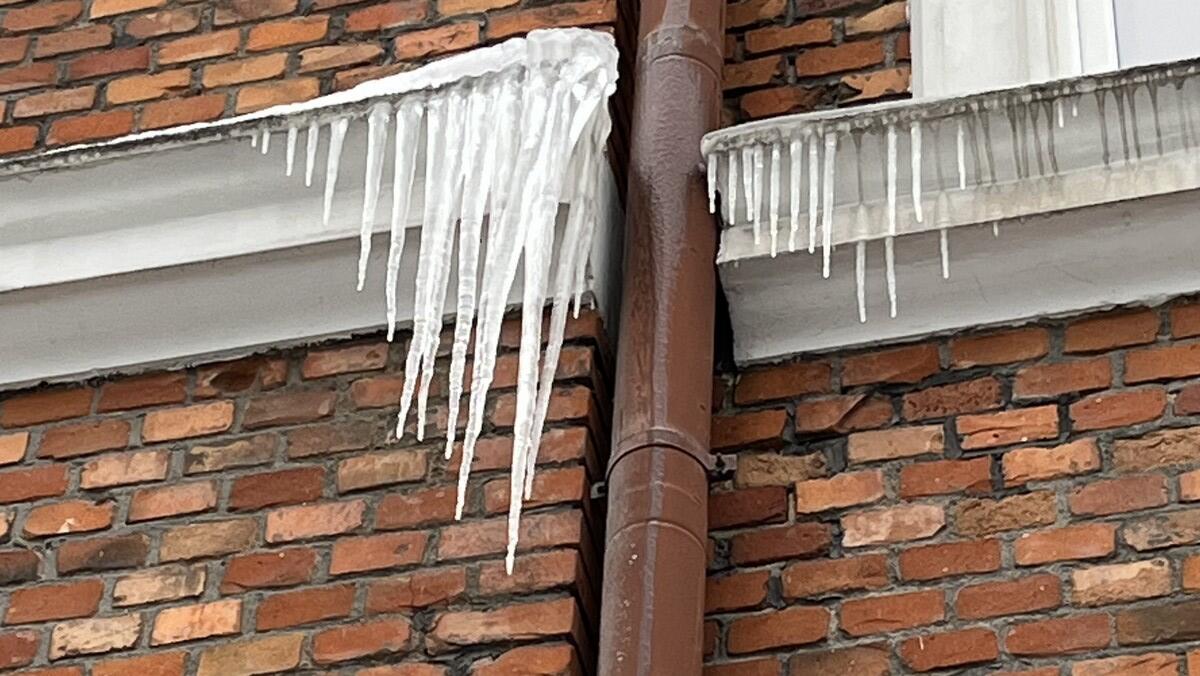 Na pierwszym zdjęciu widać fragment elewacji z cegły oraz rynnę biegnącą wzdłuż budynku. Z krawędzi dachu i elementów obróbki blacharskiej zwisają długie, ostre sople lodu. Niektóre z nich mają kilkadziesiąt centymetrów długości i tworzą gęstą linię lodowych kolców. Wygląda to na potencjalne zagrożenie dla osób przechodzących pod budynkiem.