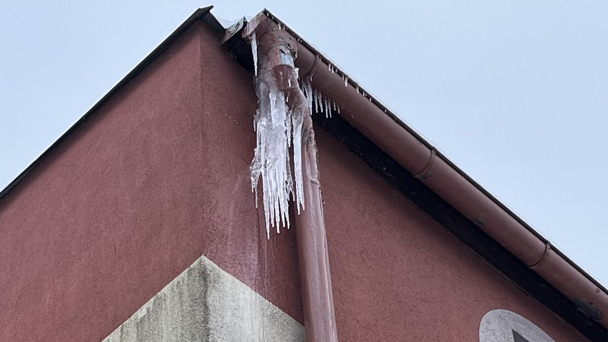 Drugie zdjęcie przedstawia narożnik dachu pokrytego powłoką w kolorze czerwonym. Z rynny oraz fragmentu obróbki dachowej wyrastają skupiska sopli lodu, zbite w dużą bryłę, z której wystają cienkie, podłużne lodowe iglice. Wygląda to jak efekt topnienia i ponownego zamarzania wody na krawędzi dachu.
