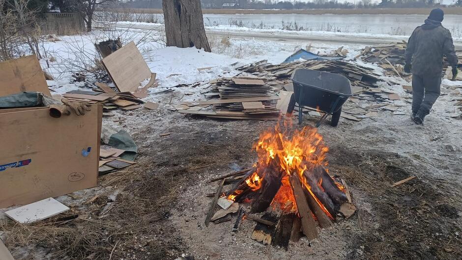 Na pierwszym zdjęciu widać zimowy krajobraz nad wodą. Na pierwszym planie pali się ognisko złożone z drewnianych desek i gałęzi – płomienie są wysokie i intensywne. Wokół ogniska leży wiele porozrzucanych kawałków drewna oraz płyt, a także taczka stojąca po prawej stronie. Po prawej stronie widoczna jest osoba ubrana w zimową odzież, odchodząca w stronę stosu desek. W tle widać zamarznięty lub zaśnieżony brzeg rzeki lub jeziora oraz drzewa bez liści.