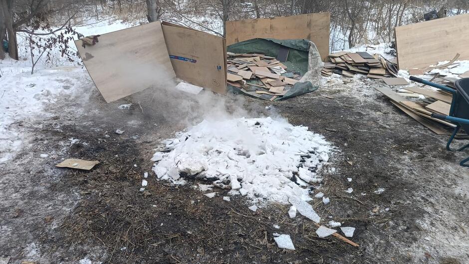 Drugie zdjęcie przedstawia to samo miejsce już po zgaszeniu ogniska. Na środku widoczna jest duża kupa śniegu i lodu, która została najprawdopodobniej użyta do ugaszenia paleniska – unoszą się z niej smugi pary. Widać również zwęglone resztki podłoża. W tle ponownie znajdują się płyty i deski ułożone oraz częściowo porozrzucane, a także prowizoryczna osłona z dużych arkuszy materiału lub kartonu oparta o drzewo, widoczna po lewej stronie.