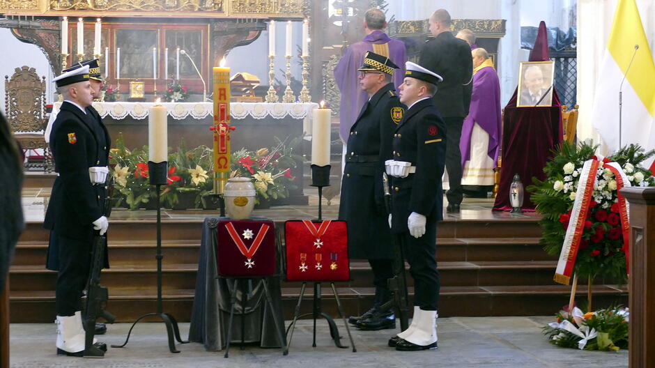 Uroczystości pogrzebowe w Bazylice Mariackiej w Gdańsku. Przy ołtarzu funkcjonariusze kompanii reprezentacyjnej Straży Miejskiej pełnią wartę honorową. Na pierwszym planie widoczna jest urna z prochami oraz wyeksponowane odznaczenia państwowe. W tle kapłani sprawujący liturgię oraz wieńce i kwiaty, które podkreślają podniosły i żałobny charakter ceremonii.