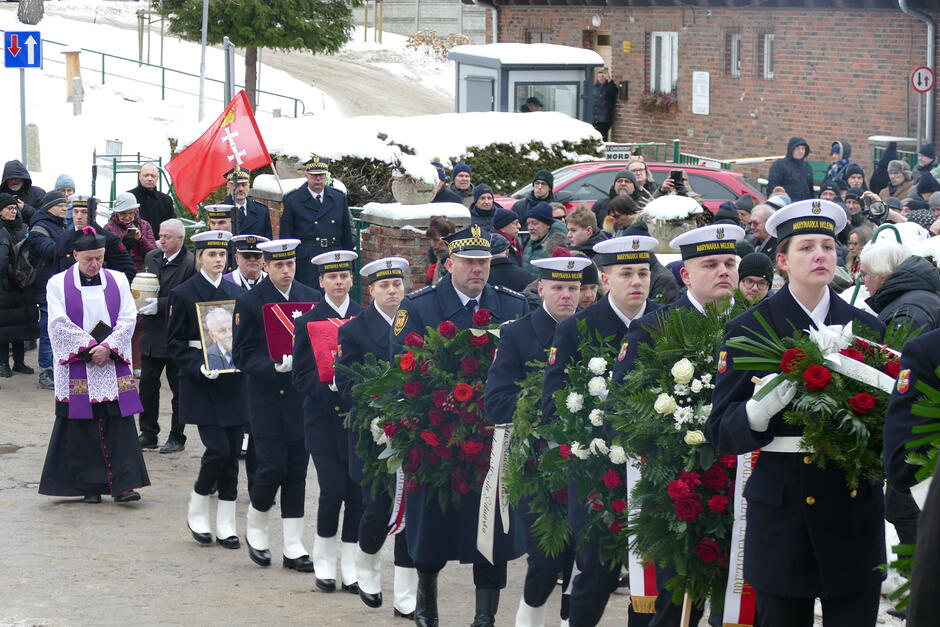 Kondukt żałobny podczas uroczystości pogrzebowych. Funkcjonariusze w galowych mundurach niosą wieńce oraz portret zmarłego, maszerując w uporządkowanym szyku. W tle widoczni są duchowni oraz licznie zgromadzeni mieszkańcy, którzy towarzyszą ceremonii, oddając hołd zmarłemu. Całość podkreśla podniosły i uroczysty charakter ostatniego pożegnania.