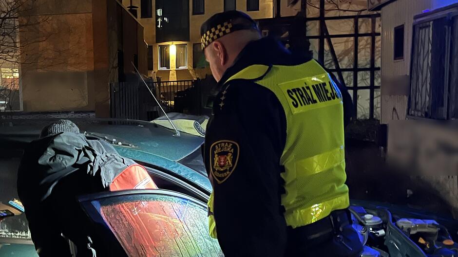 Strażnik miejski podczas nocnego patrolu sprawdza sytuację osoby siedzącej w samochodzie. Akcja odbywa się w słabo oświetlonej, miejskiej przestrzeni. Funkcjonariusz ma na sobie odblaskową kamizelkę i obserwuje wnętrze pojazdu, reagując na potencjalne zagrożenie związane z niską temperaturą.