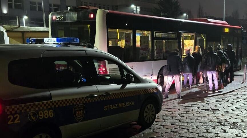Radiowóz straży miejskiej stoi na ulicy po zmroku, a w tle widać zatrzymany autobus SOS. Kilka osób wsiada do pojazdu, oświetlonego światłem wnętrza. Scena przedstawia działania służb w przestrzeni publicznej w godzinach wieczornych.