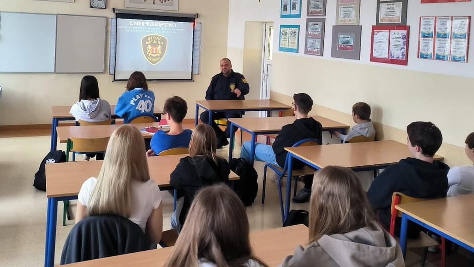 strażnik na zajęciach w klasie, przed nim siedzą uczniowie, na ekranie wyświetlana jest prezentacja