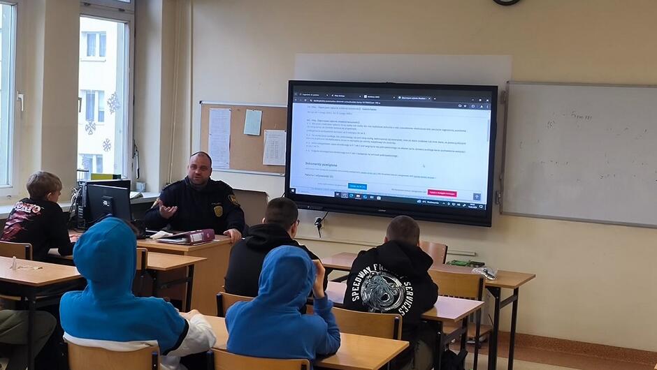 strażnik siedzi za biurkiem i prowadzi zajęcia w klasie w ławkach siedzi kliku uczniów na ekranie wyświetlana jest prezentacja. 