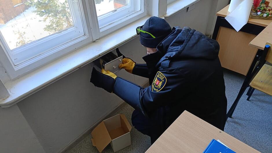 Na pierwszym zdjęciu widać strażnika miejskiego ubranego w zimową, służbową kurtkę z widocznym emblematem na ramieniu. Pracuje przy oknie wewnątrz budynku – klęczy na podłodze i ostrożnie wyciąga metalową skrzynkę z wnęki pod parapetem. Na dłoniach ma grube, ochronne rękawice. Obok na podłodze stoi kartonowe pudełko, prawdopodobnie przygotowane do przeniesienia zawartości skrzynki.