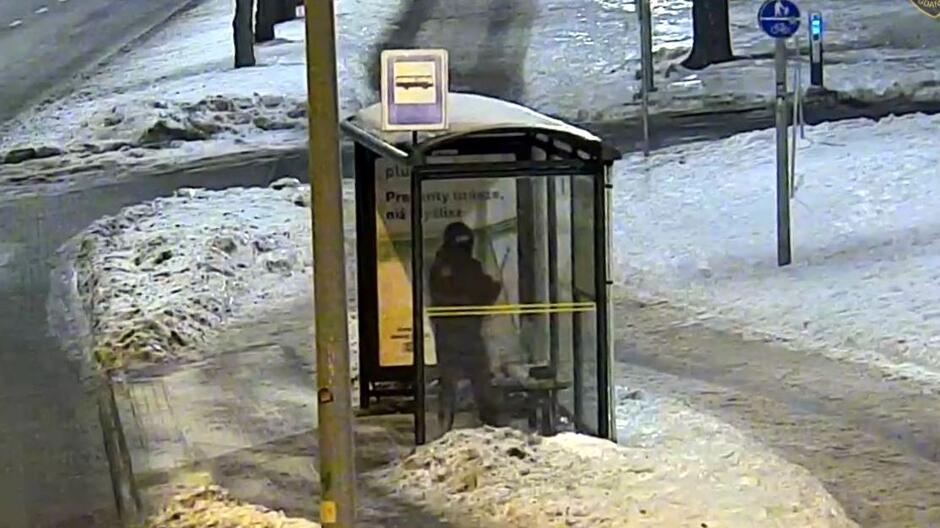 Na zdjęciu widać oszkloną wiatę przystanku autobusowego stojącą przy zaśnieżonej ulicy. W środku znajduje się pojedyncza osoba, która trzyma rękę uniesioną w kierunku szyby. Wokół przystanku leży śnieg, a chodniki i droga są oświetlone ulicznymi lampami. W tle widoczne są znaki drogowe oraz pusty chodnik.