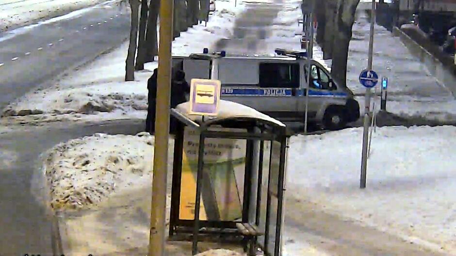 Na zdjęciu widać ośnieżony przystanek autobusowy stojący przy ulicy. Przed przystankiem zatrzymał się policyjny radiowóz w biało‑niebieskich barwach. Obok pojazdu stoi funkcjonariusz. Ulica i chodniki są pokryte śniegiem, a otoczenie jest oświetlone latarniami. Scena wygląda na ujęcie z monitoringu miejskiego wykonane nocą lub wczesnym rankiem.