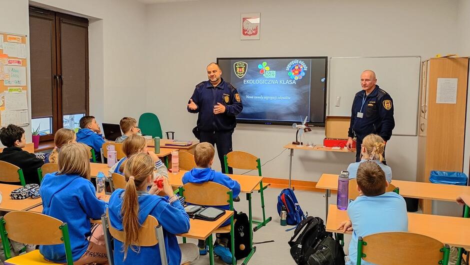 W jasnej, nowoczesnej sali lekcyjnej odbywa się spotkanie edukacyjne prowadzone przez dwóch funkcjonariuszy straży miejskiej. Stoją oni na tle dużego monitora, na którym wyświetlona jest prezentacja pt. „Ekologia w klasie”. Uczniowie w niebieskich bluzach siedzą w kilku rzędach przy szkolnych ławkach, uważnie słuchając prowadzących. Na tablicach i ścianach widoczne są notatki oraz szkolne ogłoszenia, a na ławkach znajdują się przybory szkolne i bidony. Atmosfera wskazuje na angażujące zajęcia o charakterze edukacyjno‑profilaktycznym.